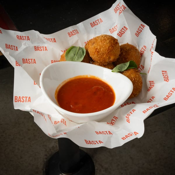 Golden-brown arancini with fresh basil and a side of red marinara sauce in BASTA-branded paper.