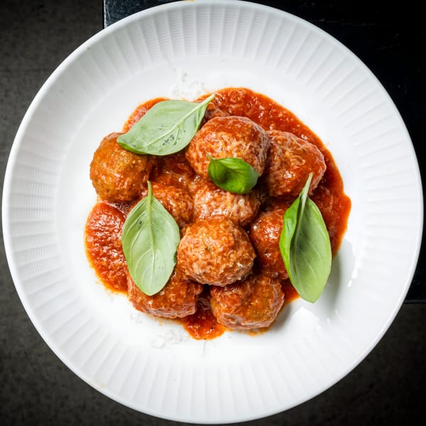 BASTA Pasteria's Italian meatballs in rich tomato sauce, garnished with fresh basil leaves on a white plate.