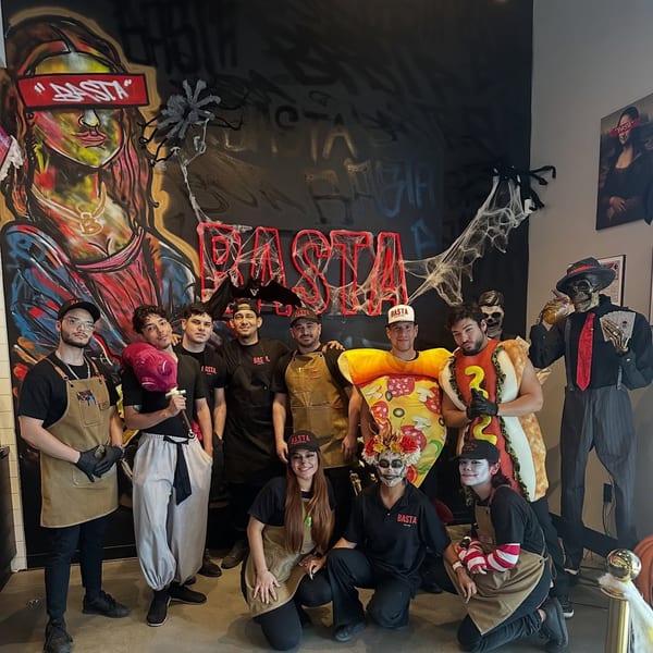 BASTA Pasteria staff in Halloween costumes pose in front of a graffiti wall with spiderwebs.