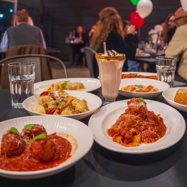 Table laden with various Italian pasta dishes, a milkshake, and water glasses, with blurred diners in the background.